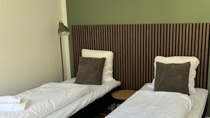 Zwei Einzelbetten mit weißer Bettwäsche, ein Holzschrankbrettchen am Kopfteil, ein kleiner runder Tisch und eine Stehlampe.