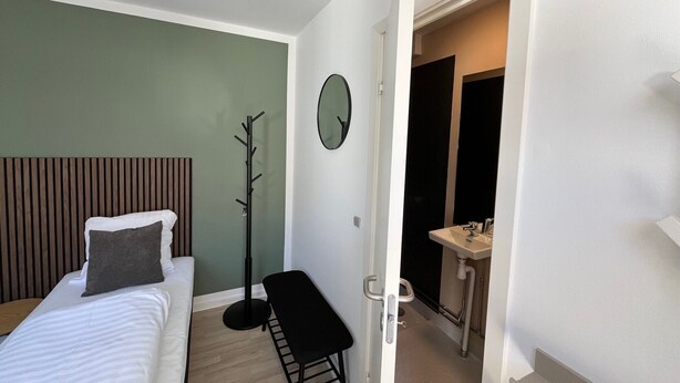 Ein Schlafzimmer mit Bett, Bank, Kleiderständer, Wanduhr und einem durchgegangenen Türrahmen erreichbarem Badezimmer.