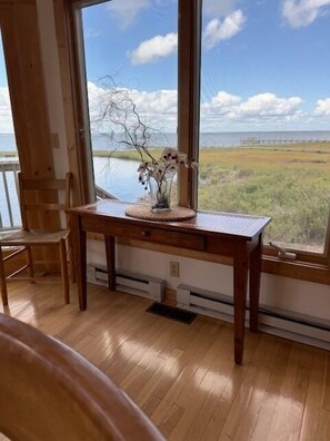 A wooden desk with a vase of flowers, a chair, and a view of a body of water and grassy landscape through large windows. - Sialang- Private Waterfront Pier, Hot Tub, Kayaks (Chincoteague Island)
