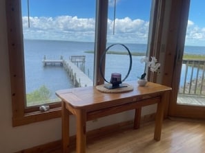 A wooden table with a candle and a small plant by a window overlooking a body of water and a dock. - Sialang- Private Waterfront Pier, Hot Tub, Kayaks (Chincoteague Island)