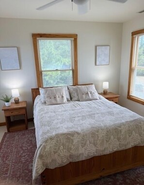 Une chambre à coucher avec un grand lit, deux tables de chevet, un ventilateur de plafond et deux fenêtres équipées de stores.