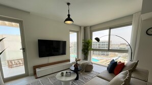 A modern living room with a flat-screen TV, a round coffee table, a sofa with cushions, a potted plant, and a view of a swimming pool and buildings. - 2 BR Sapphire 405 in Marassi North Coast (El Alamein)