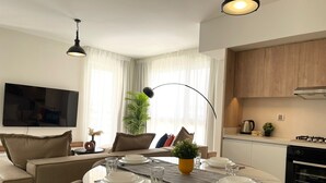 A modern living room with a dining table set for two, a sofa, a TV mounted on the wall, and a kitchen area with cabinets and appliances. - 2 BR Sapphire 405 in Marassi North Coast (El Alamein)