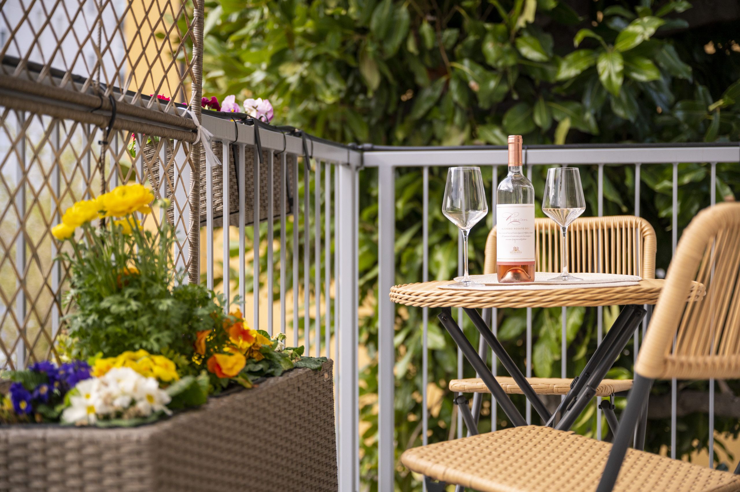 A wicker table with a bottle and glasses, a wicker chair, and a flower arrangement on a balcony.