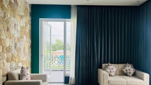 A room with a stone wall, a beige sofa, a dark curtain, and a view through a glass door. - Mastiff Citadel RC (Vikasnagar)