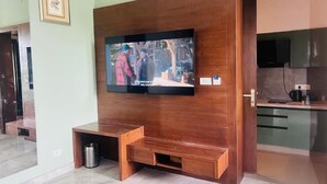 A living room with a wooden entertainment unit, a television, and a kitchen area. - Mastiff Citadel RC (Vikasnagar)