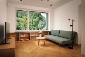Ein modernes Wohnzimmer mit einem großen Fenster, Holzboden, einem grünen Sofa, einem kleinen Tisch und einer Stehlampe.