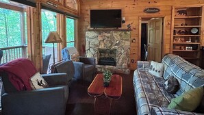 Interior de una cabaña de madera con chimenea de piedra, televisión y estantería.