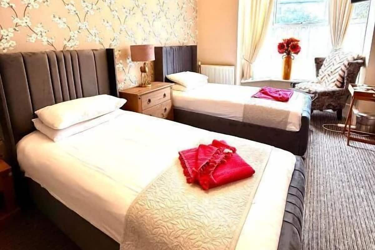 A hotel room with two beds, a window with curtains, a small table, and a wall with a floral pattern.