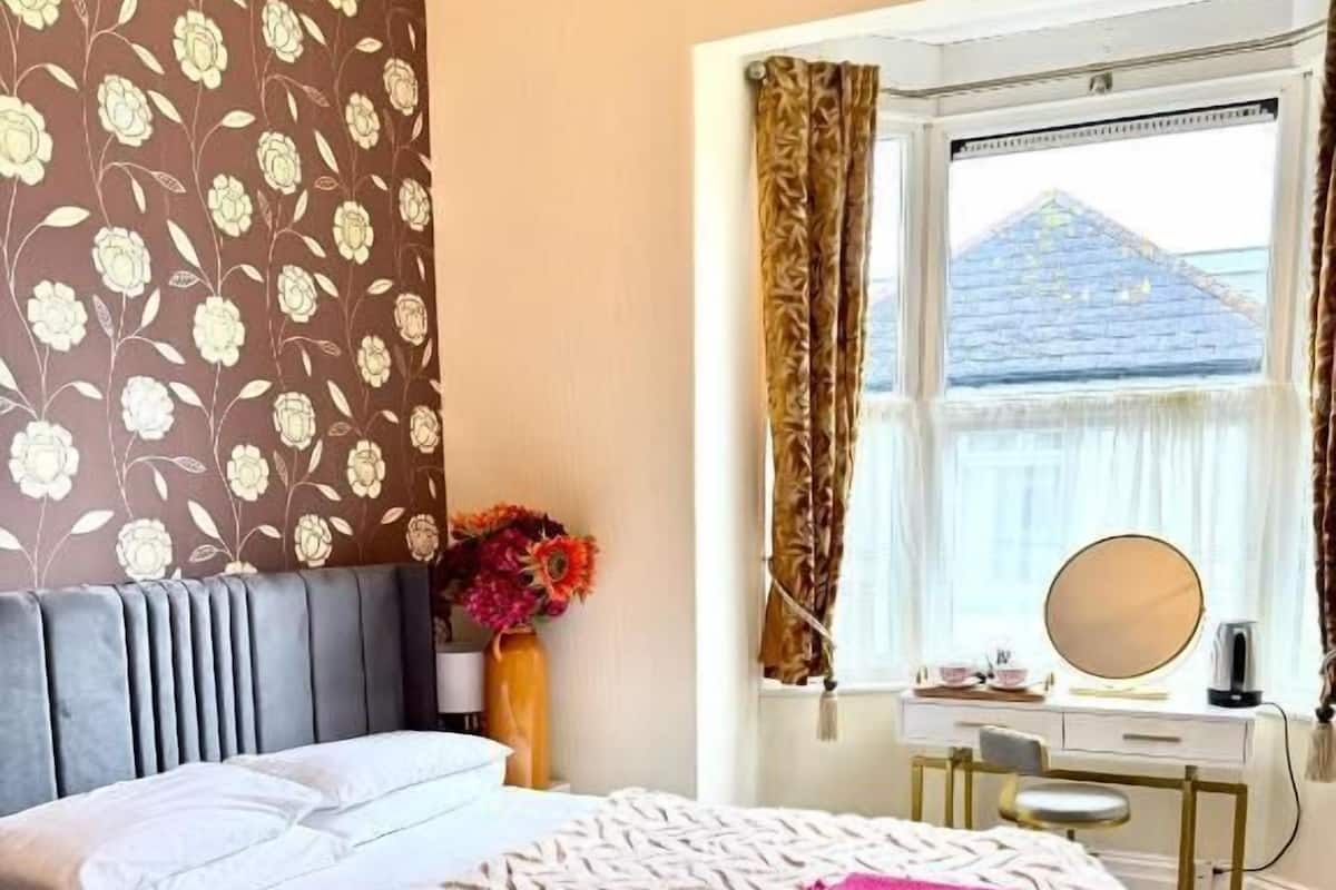 A neatly made bed with a patterned bedspread, a bedside table with a mirror, and a window with curtains.