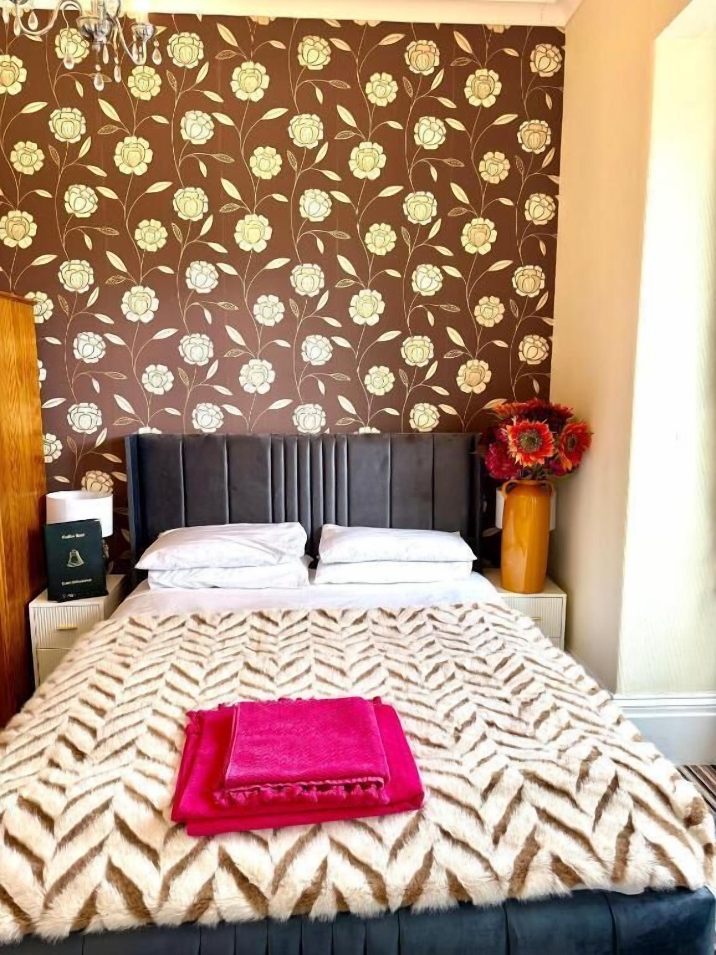 A bedroom with a patterned wallpaper, a bed with a white and brown bedspread, a red throw, and a vase with flowers.