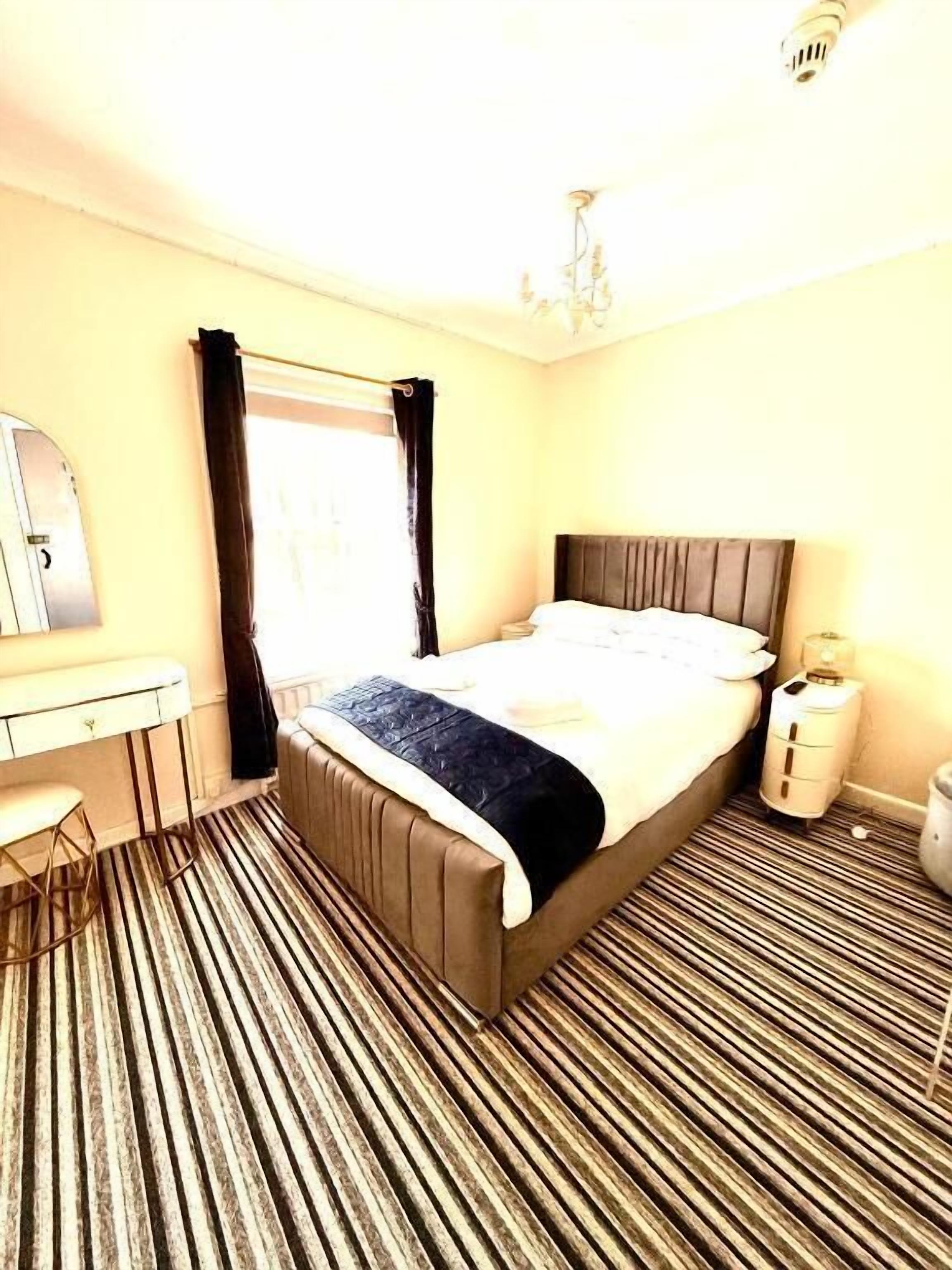 A hotel room with a bed, a desk, a chair, a mirror, a window with curtains, and a radiator.