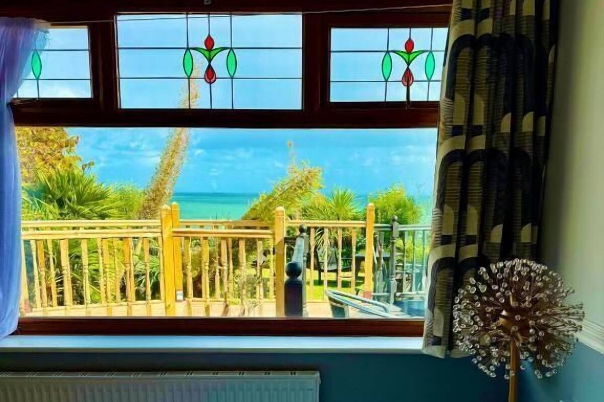 A room with a bed, a view of the sea through a window, and a balcony with a railing.