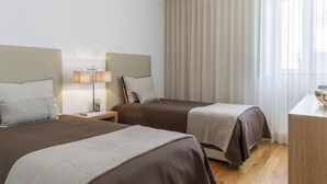 Ett hotellrum med två sängar, ett nattduksbord med en lampa, ett skrivbord och ett stort fönster med gardiner.
