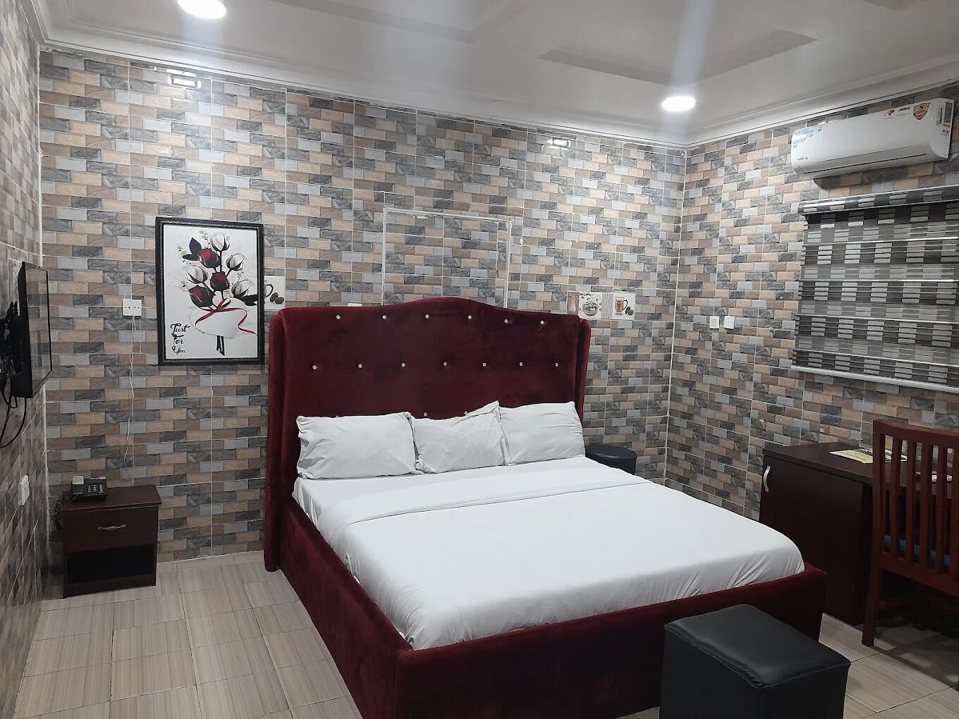 A bedroom with a red upholstered bed, white bedding, a small bedside table, a wall-mounted air conditioner, and a framed picture on the wall.
