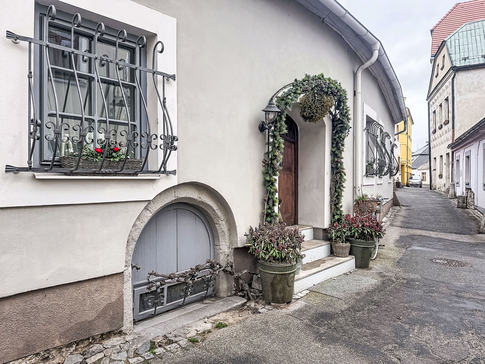 A narrow street with a building featuring a window with metal bars, a wooden door, and a small porch area with potted plants.