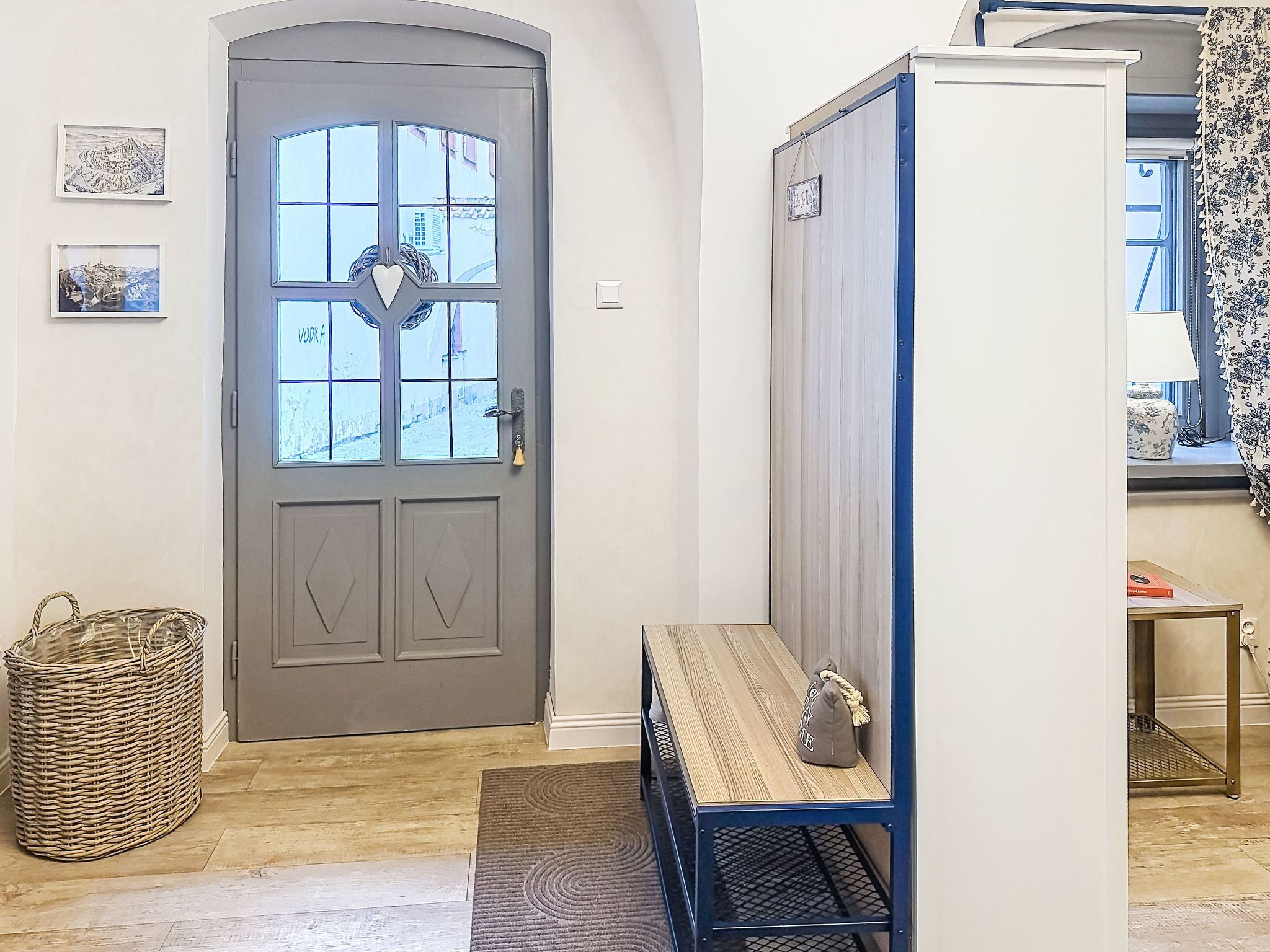 A room with a grey door, a wicker basket, a bench with a bag, and a window with a curtain.