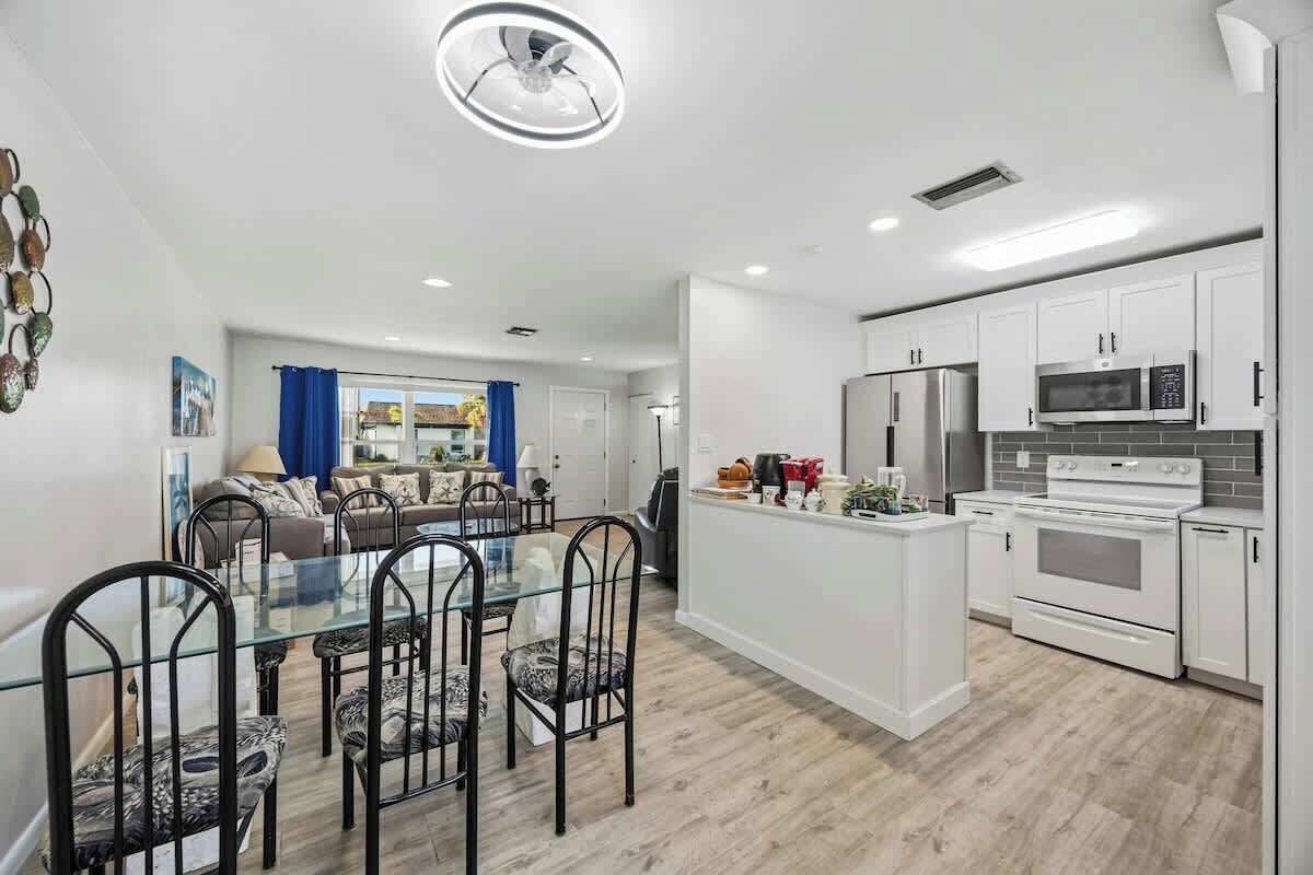 Uma cozinha moderna com armários brancos, uma ilha central e uma área de jantar com mesa de vidro e cadeiras de metal.