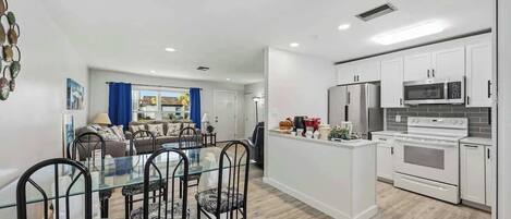 Uma cozinha moderna com armários brancos, uma ilha central e uma área de jantar com mesa de vidro e cadeiras de metal.