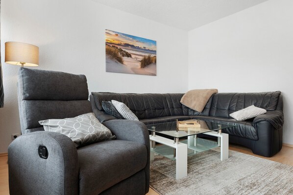 A modern living room with a grey armchair, a black leather sofa, a glass coffee table, and a wall-mounted picture of a beach scene.