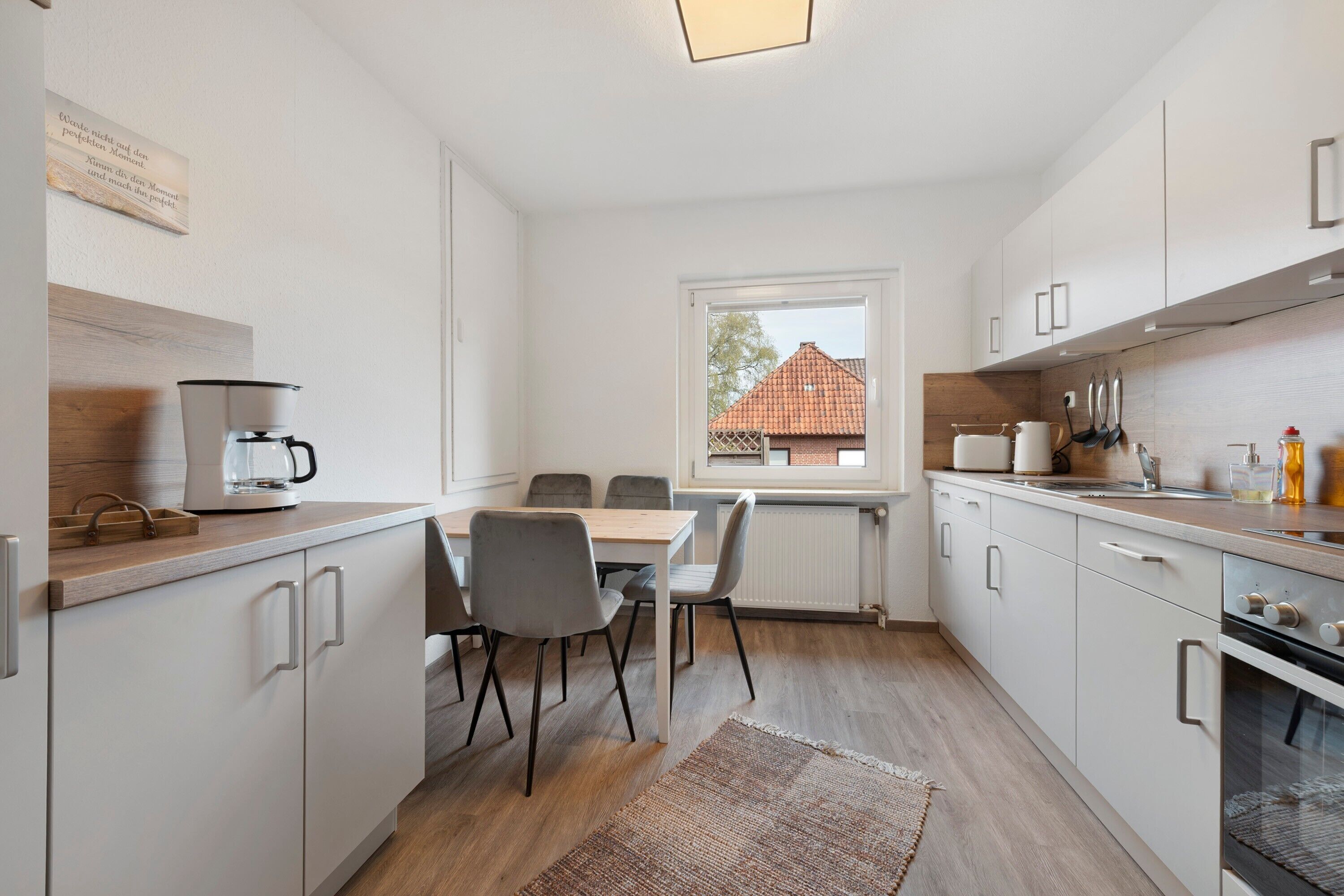 Una cucina moderna con mobili bianchi, un tavolo da pranzo con sedie, una macchina per il caffè e una finestra con vista su una casa.