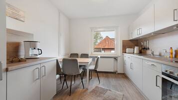 Una cucina moderna con mobili bianchi, un tavolo da pranzo con sedie, una macchina per il caffè e una finestra con vista su una casa.
