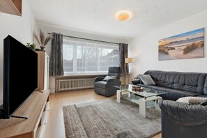 A modern living room with a flat-screen TV, a sofa, a coffee table, and a large window with curtains.