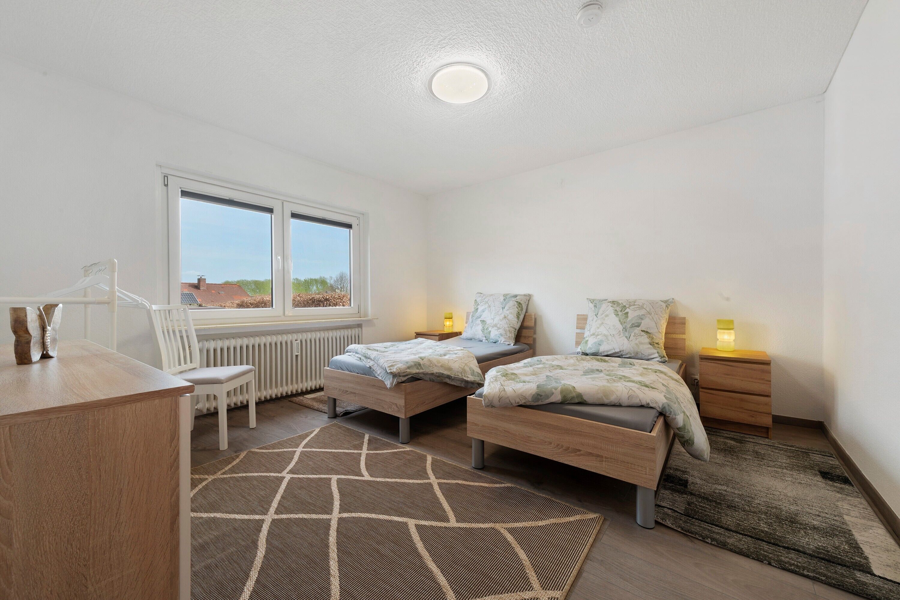 Une chambre avec deux lits, un bureau, une chaise, un radiateur et une vue sur l’extérieur.