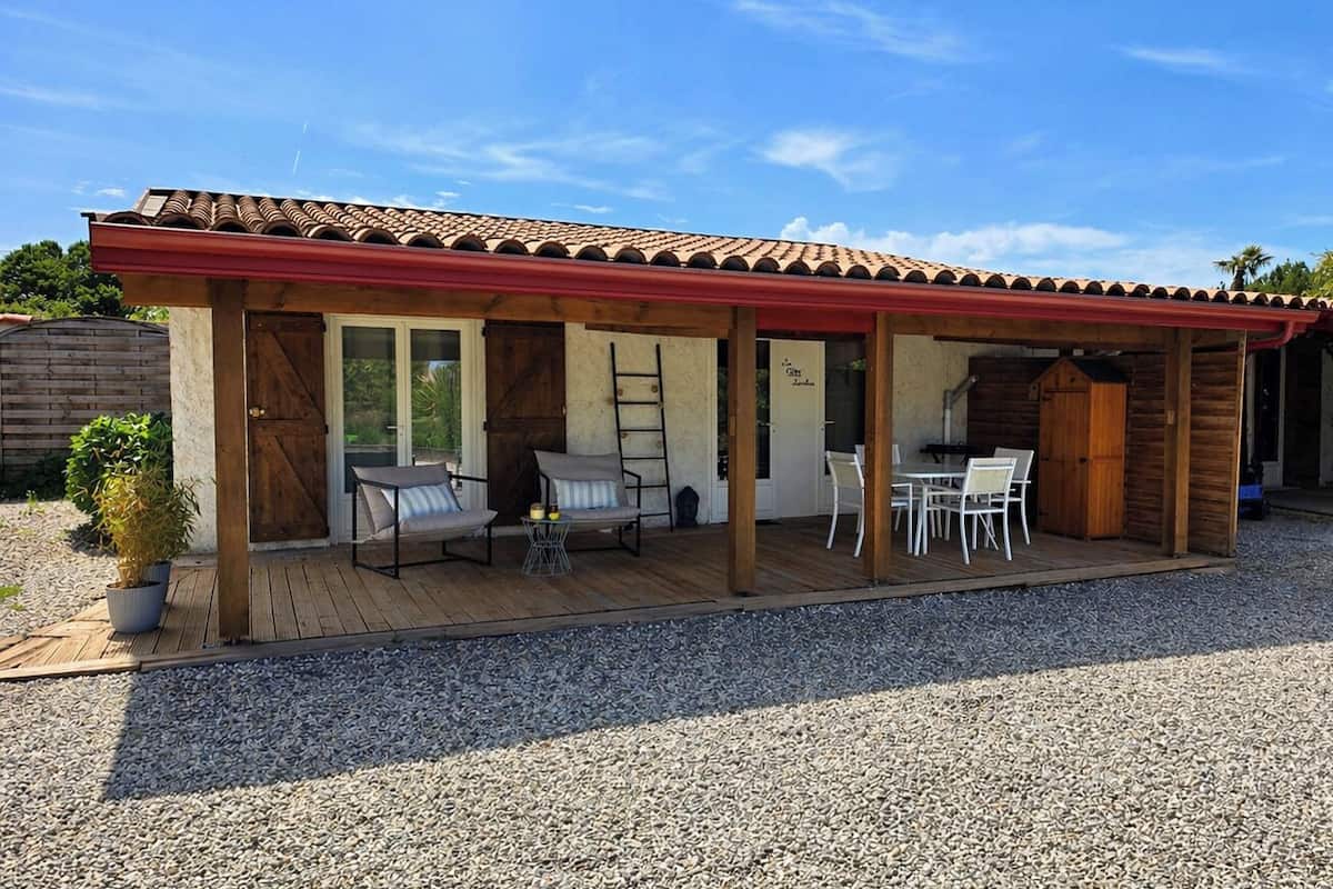Een overdekt terras buiten, met houten vloer, een tafel, stoelen en een potplant.