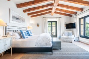 Ein Schlafzimmer mit einem Bett, Nachttischen, einem Sessel, einem Fernseher und einem Fenster mit Vorhängen.