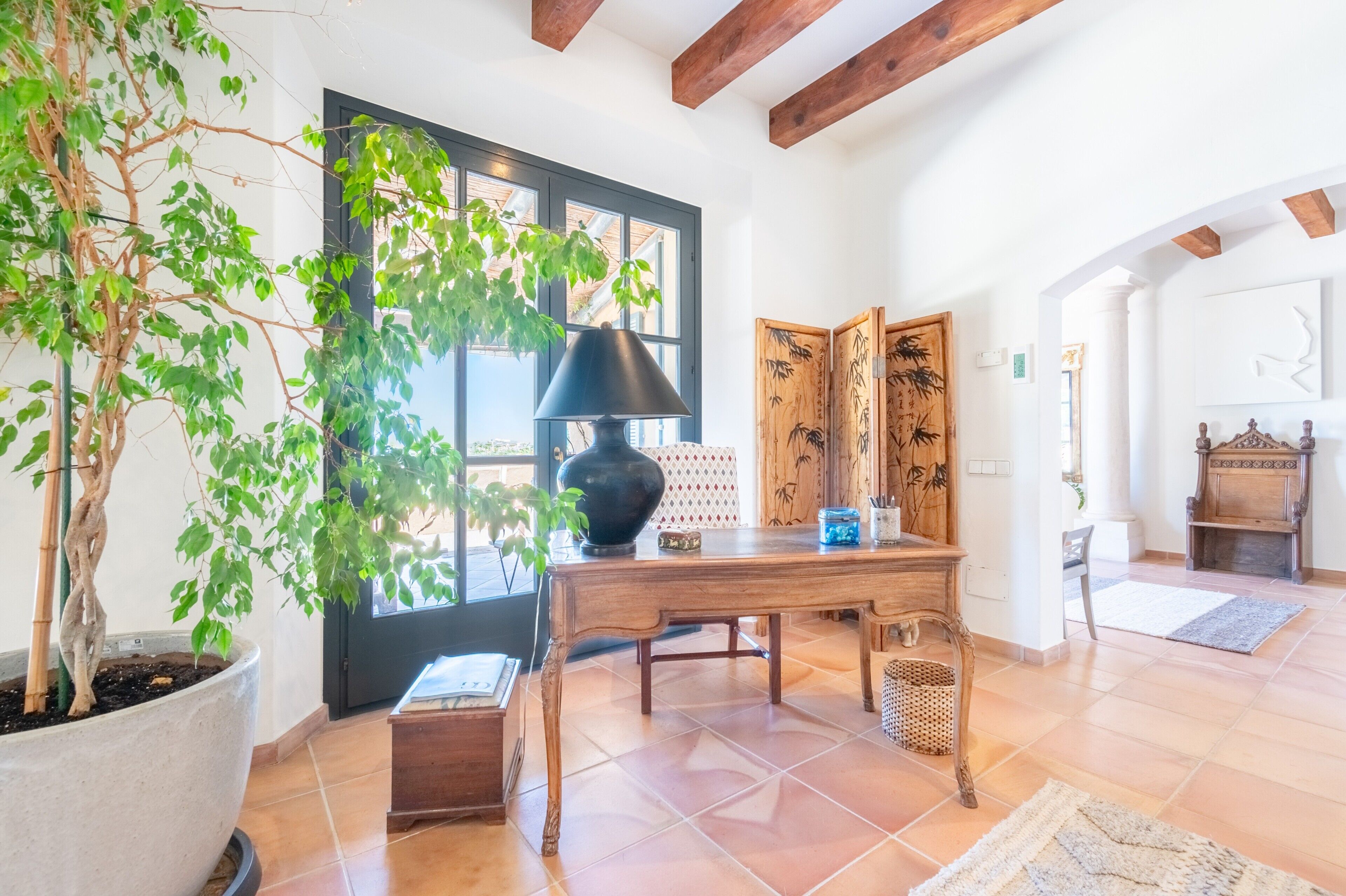 Une pièce avec une grande plante en pot, une table en bois surmontée d’une lampe noire, un paravent japonais traditionnel et une vue par la fenêtre.