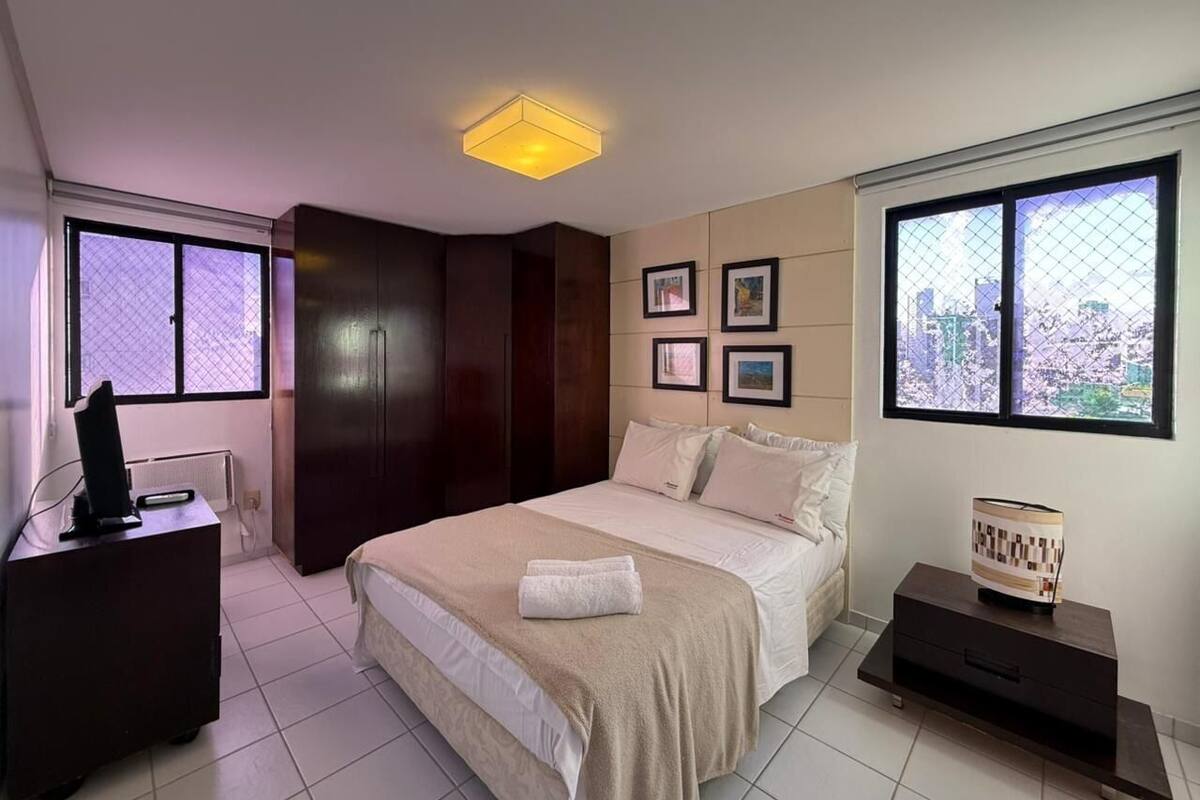 A bedroom with a bed, a desk, a dresser, a nightstand, and a window with a city view.