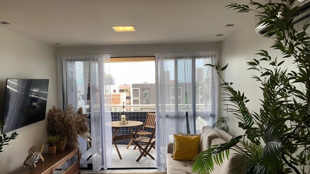En stue med sofa, fjernsyn, spisebord med stole og en balkon med udsigt til bygninger.