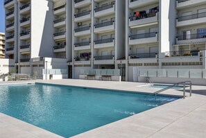 Ein Schwimmbecken mit Metallleiter, umgeben von einem Gebäude mit mehreren Balkonen und einer flachen Dachterrasse.