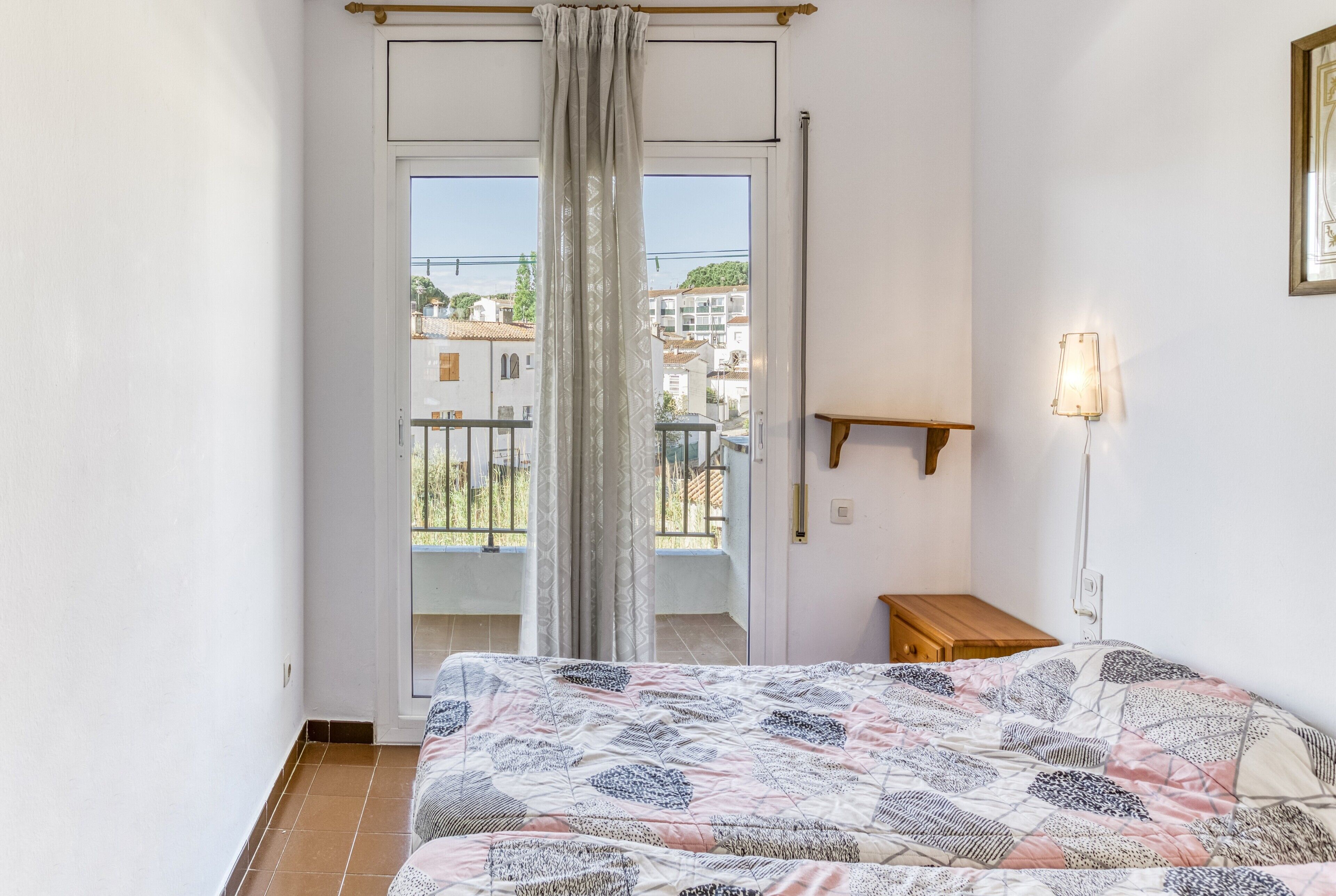 A bedroom with a bed, a wooden nightstand, a wall-mounted lamp, and a balcony door.