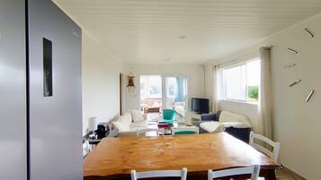 A dining area with a wooden table and white chairs, a television, and a view of the outdoors through a window.