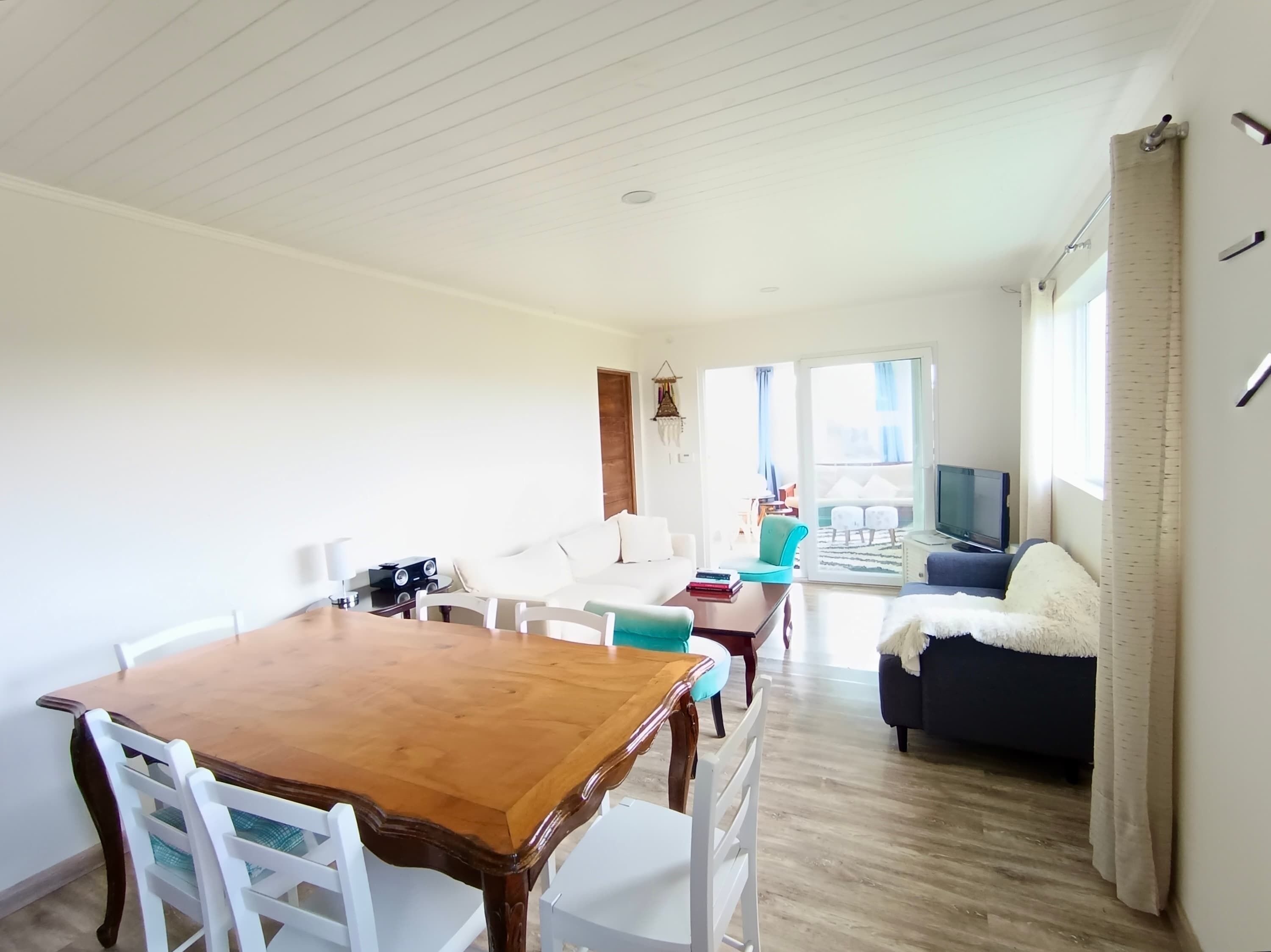A spacious living area with a wooden dining table, white chairs, a sofa, and a television.