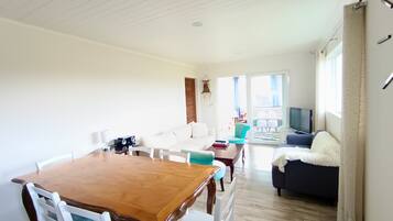 A spacious living area with a wooden dining table, white chairs, a sofa, and a television.