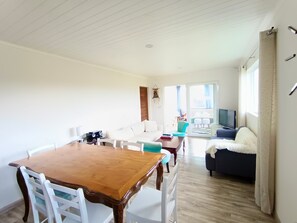A spacious living area with a wooden dining table, white chairs, a sofa, and a television.