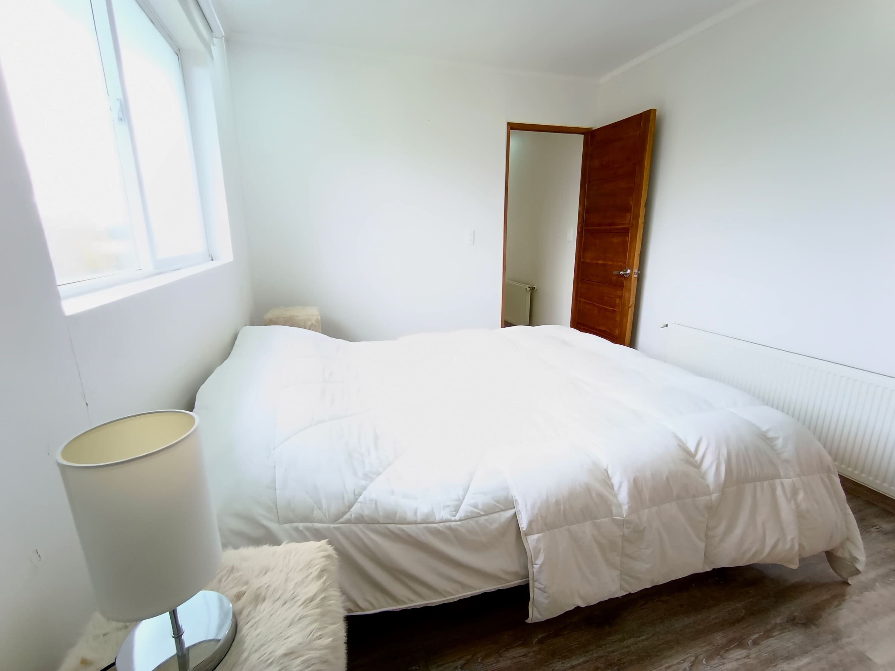 A bedroom with a bed, a bedside lamp, a wooden door, and a window.