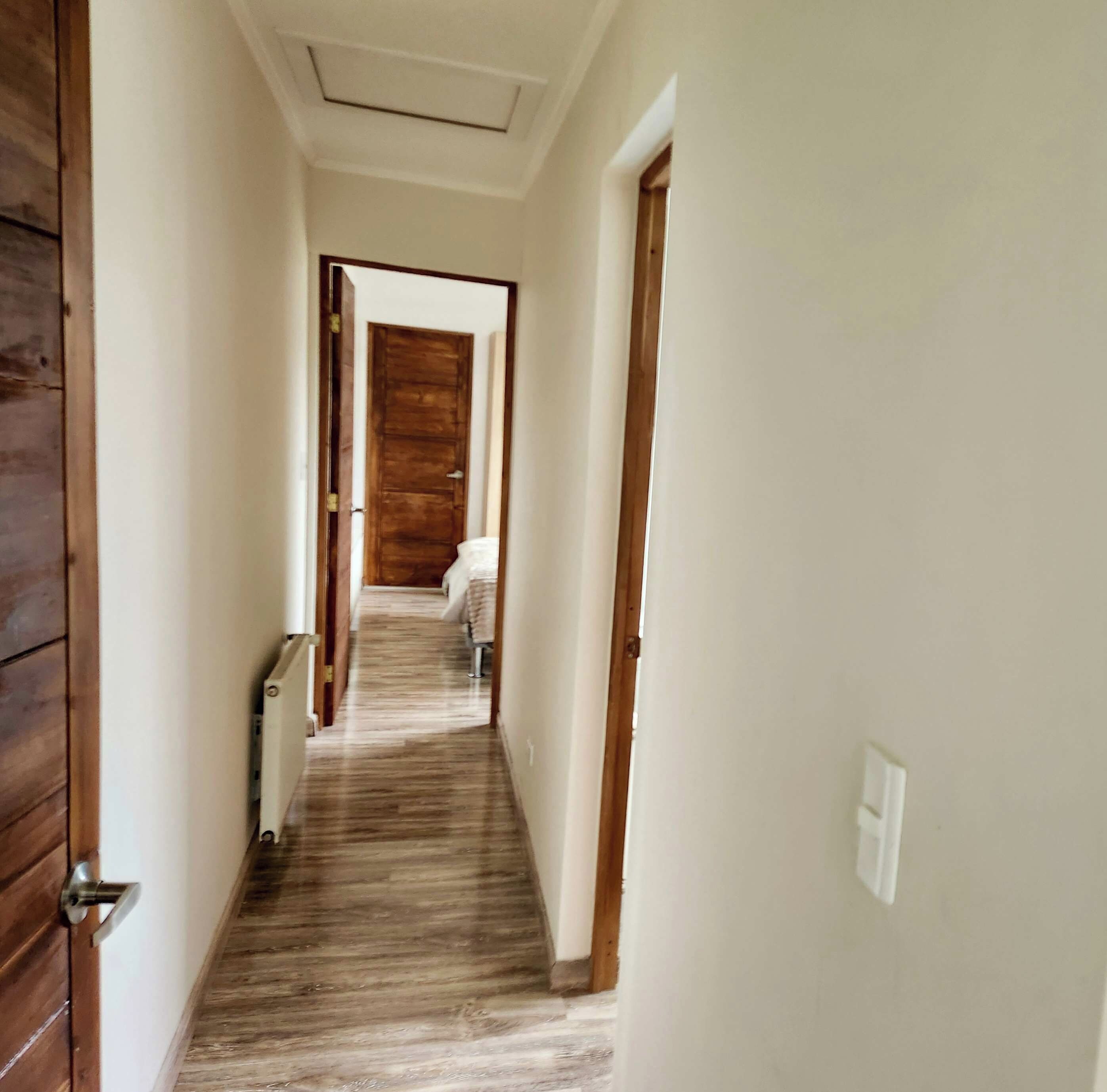 A narrow hallway with wooden flooring and white walls, leading to a room with a bed and a window.