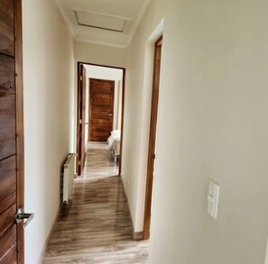 A narrow hallway with wooden flooring and white walls, leading to a room with a bed and a window.