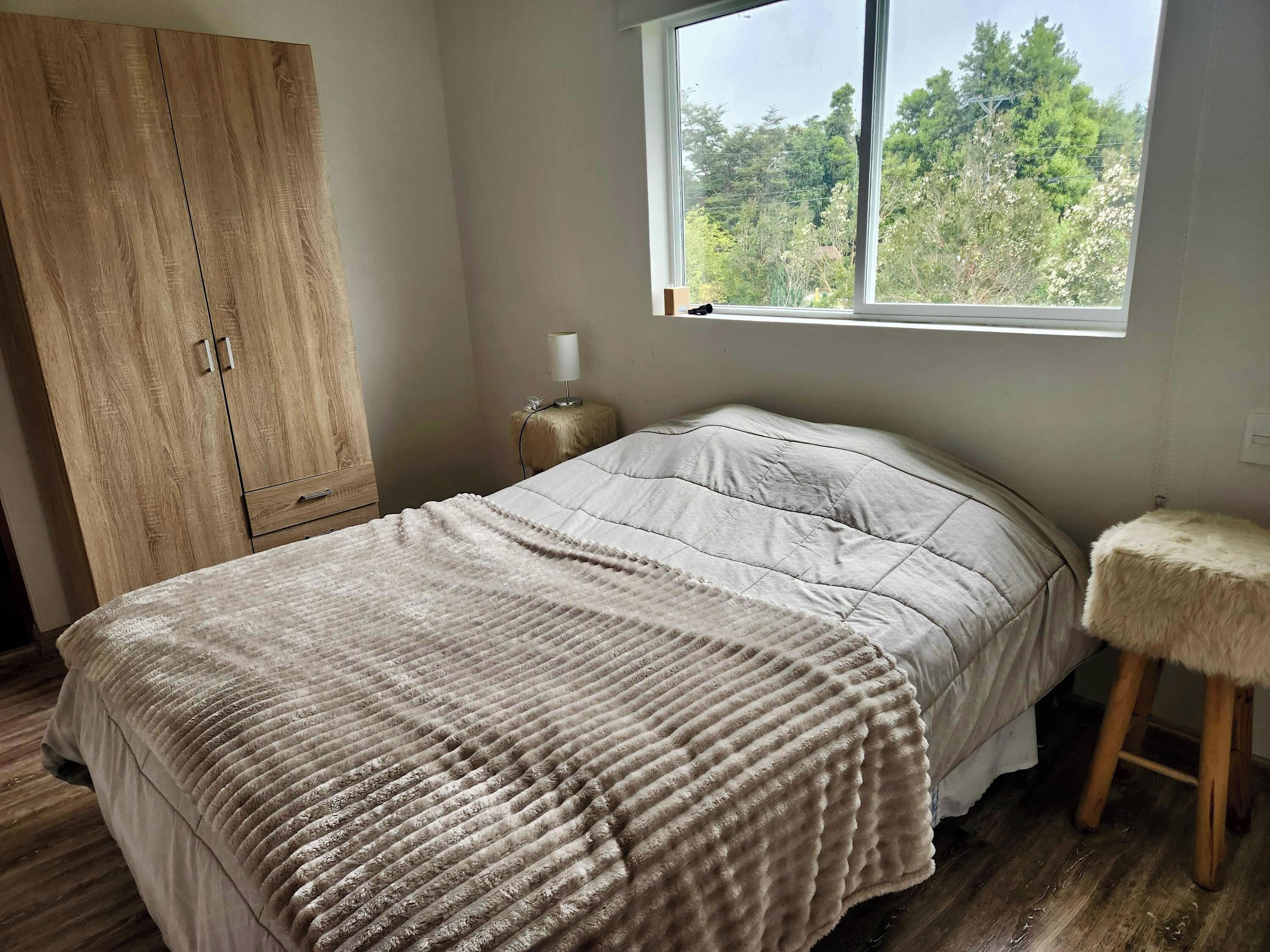 A bedroom with a bed, a wooden wardrobe, a nightstand with a lamp, a window with a view of trees, and a small stool.