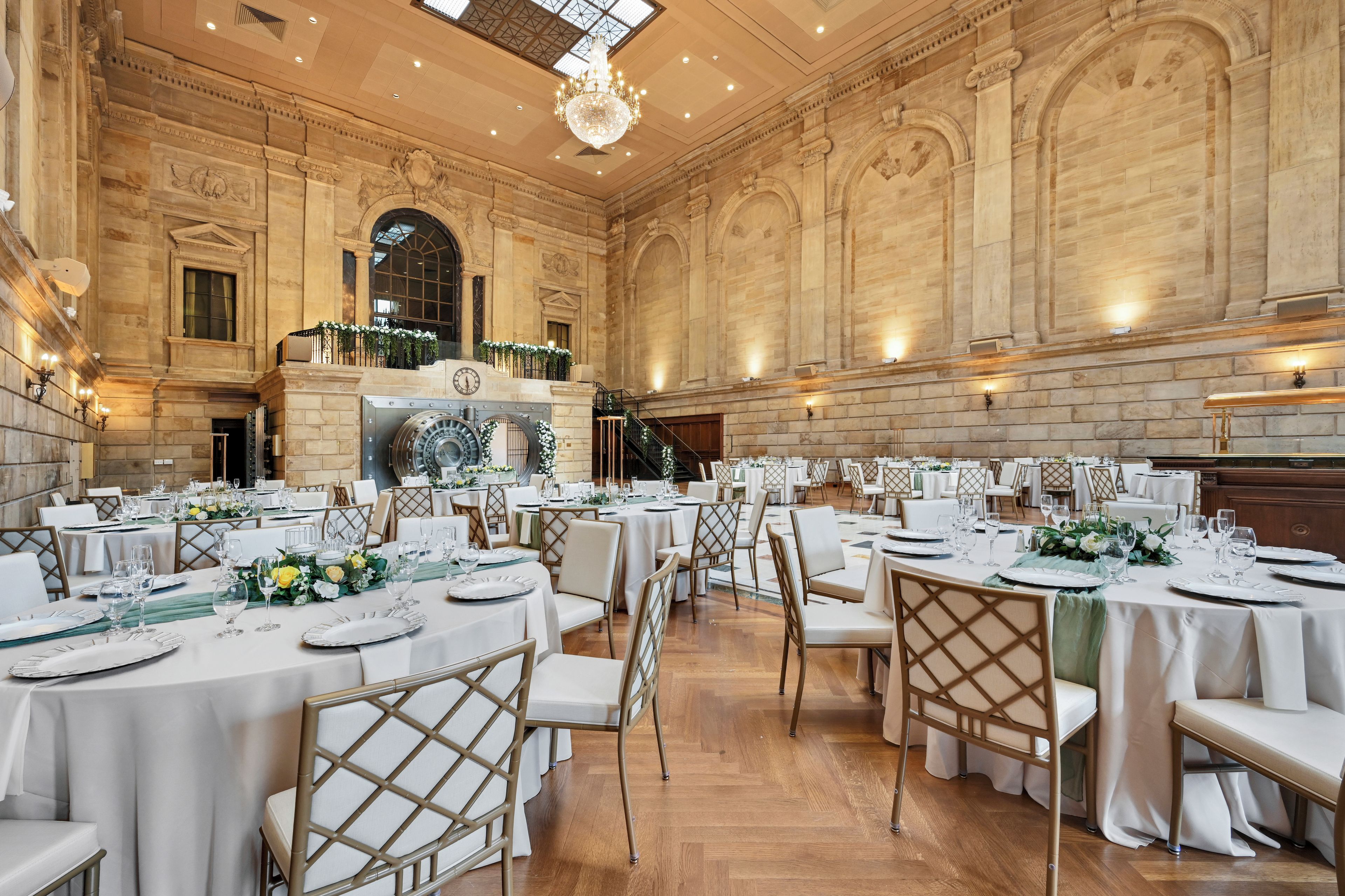 A grand banquet hall with high ceilings, ornate stone walls, and a large chandelier. Tables are set with white tablecloths and gold chairs.