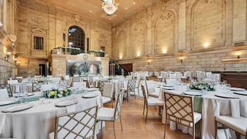 A grand banquet hall with high ceilings, ornate stone walls, and a large chandelier. Tables are set with white tablecloths and gold chairs.