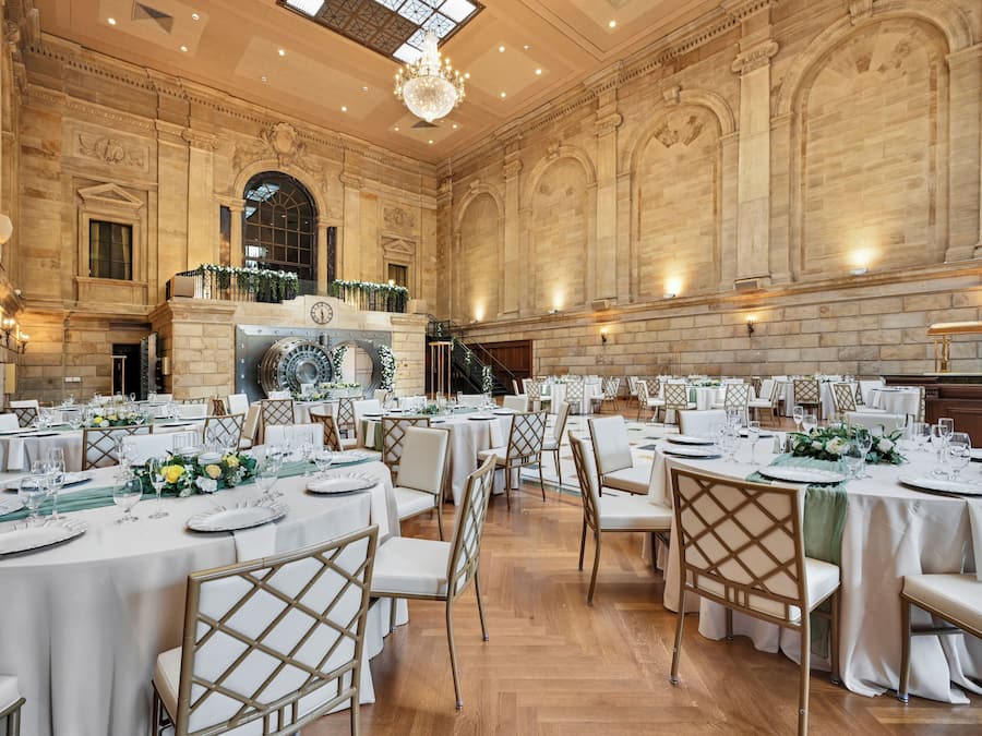 A grand banquet hall with high ceilings, ornate stone walls, and a large chandelier. Tables are set with white tablecloths and gold chairs.
