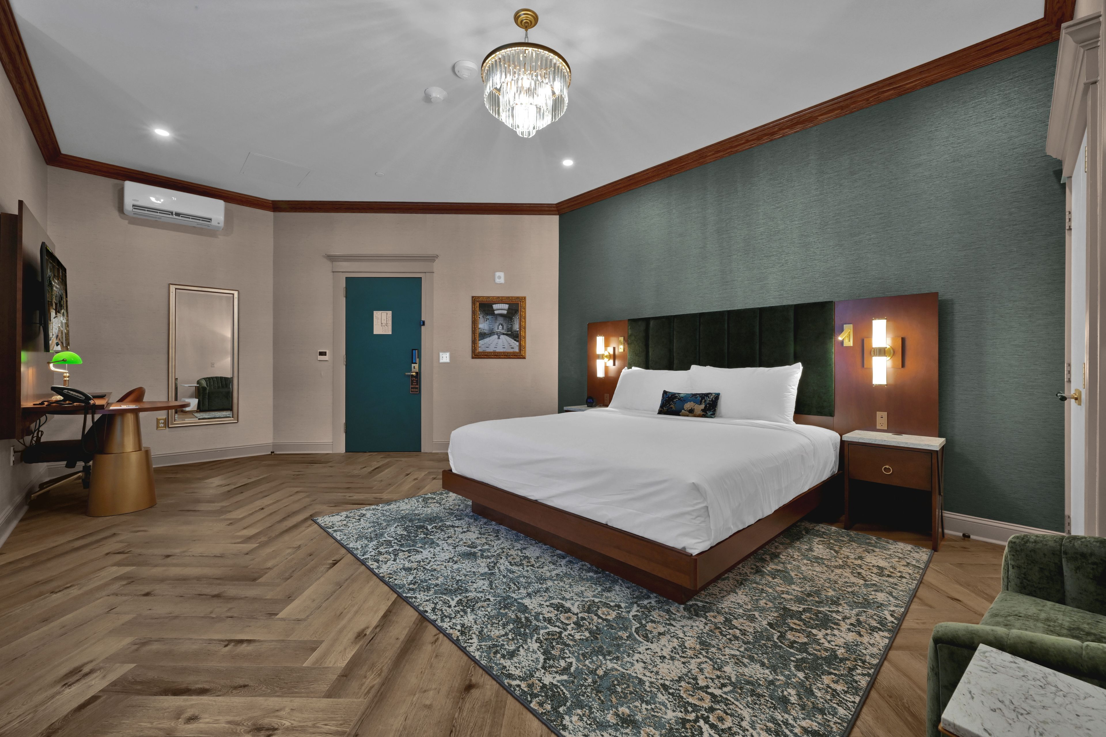 A hotel room with a large bed, a desk, a chair, a desk lamp, a wall-mounted air conditioner, a chandelier, and a door.