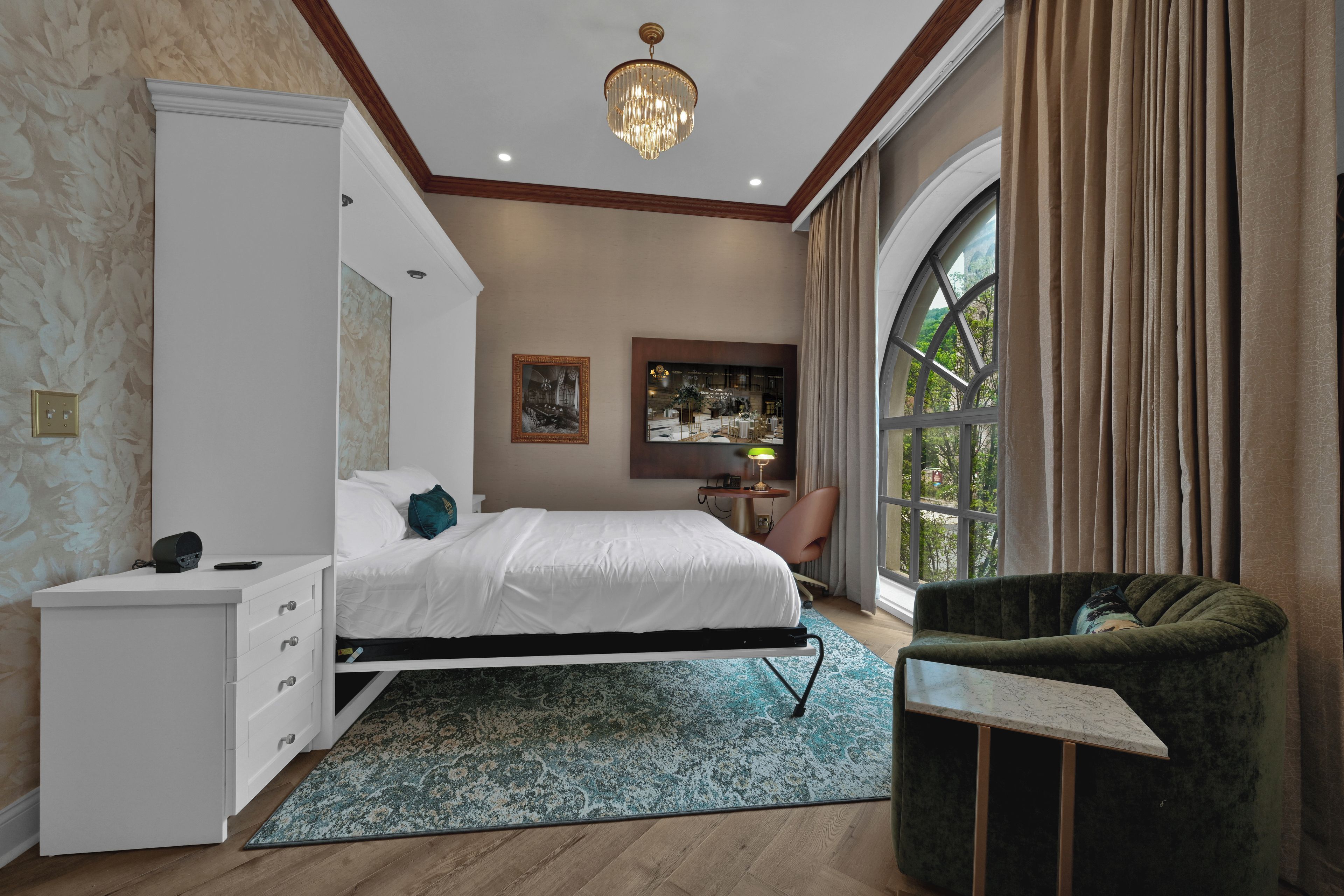 Camera d'albergo con un letto, una scrivania, una sedia, un'ampia finestra e un lampadario.