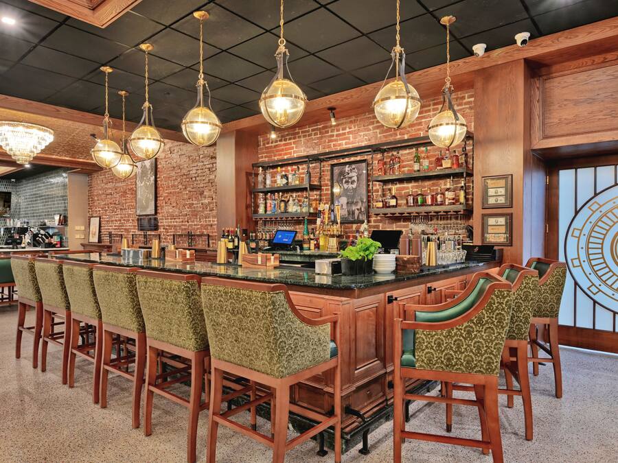 A bar area with a long counter, multiple bar stools, and a brick wall backdrop.