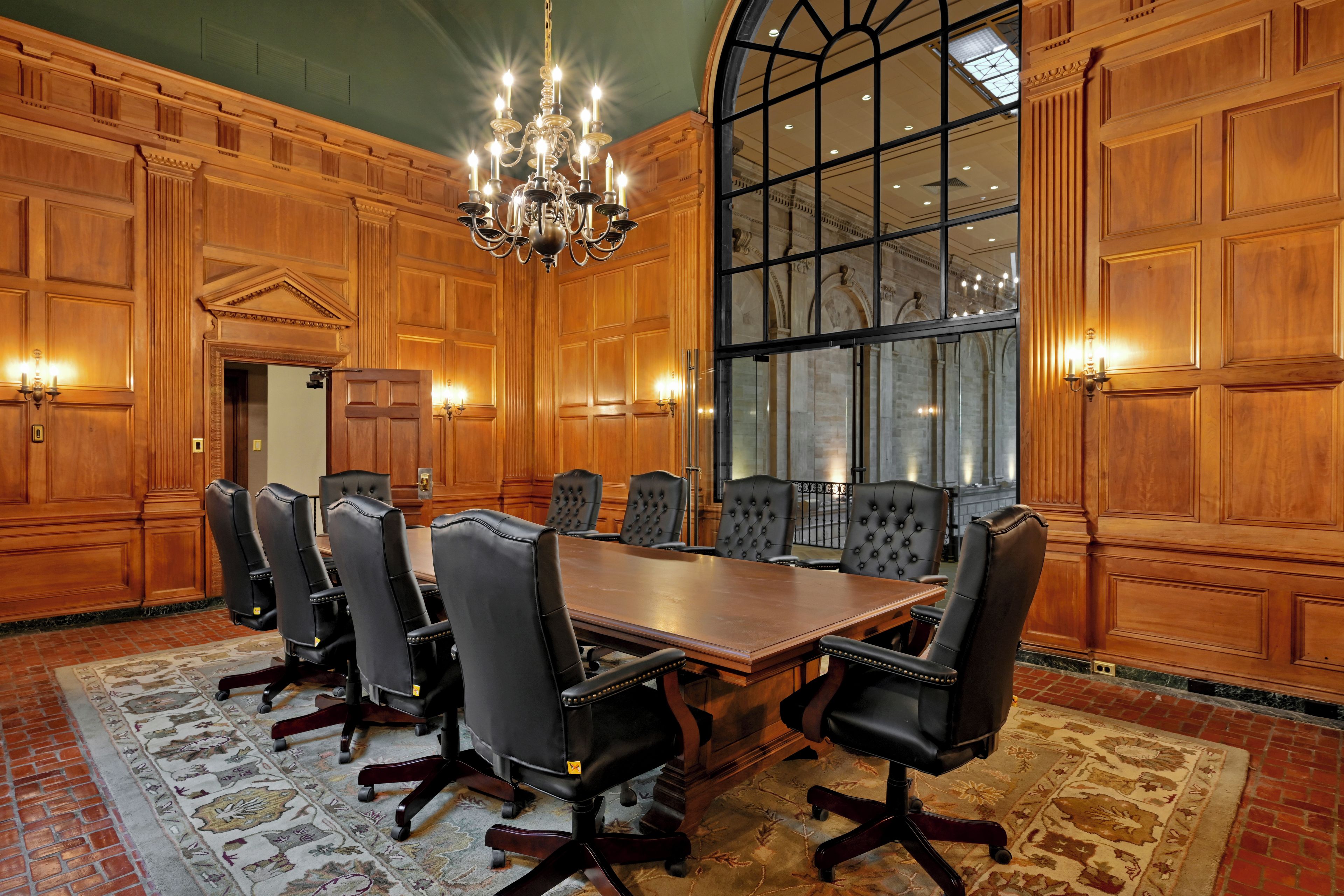 A conference room with a large wooden table, black leather chairs, a chandelier, and a large arched window.